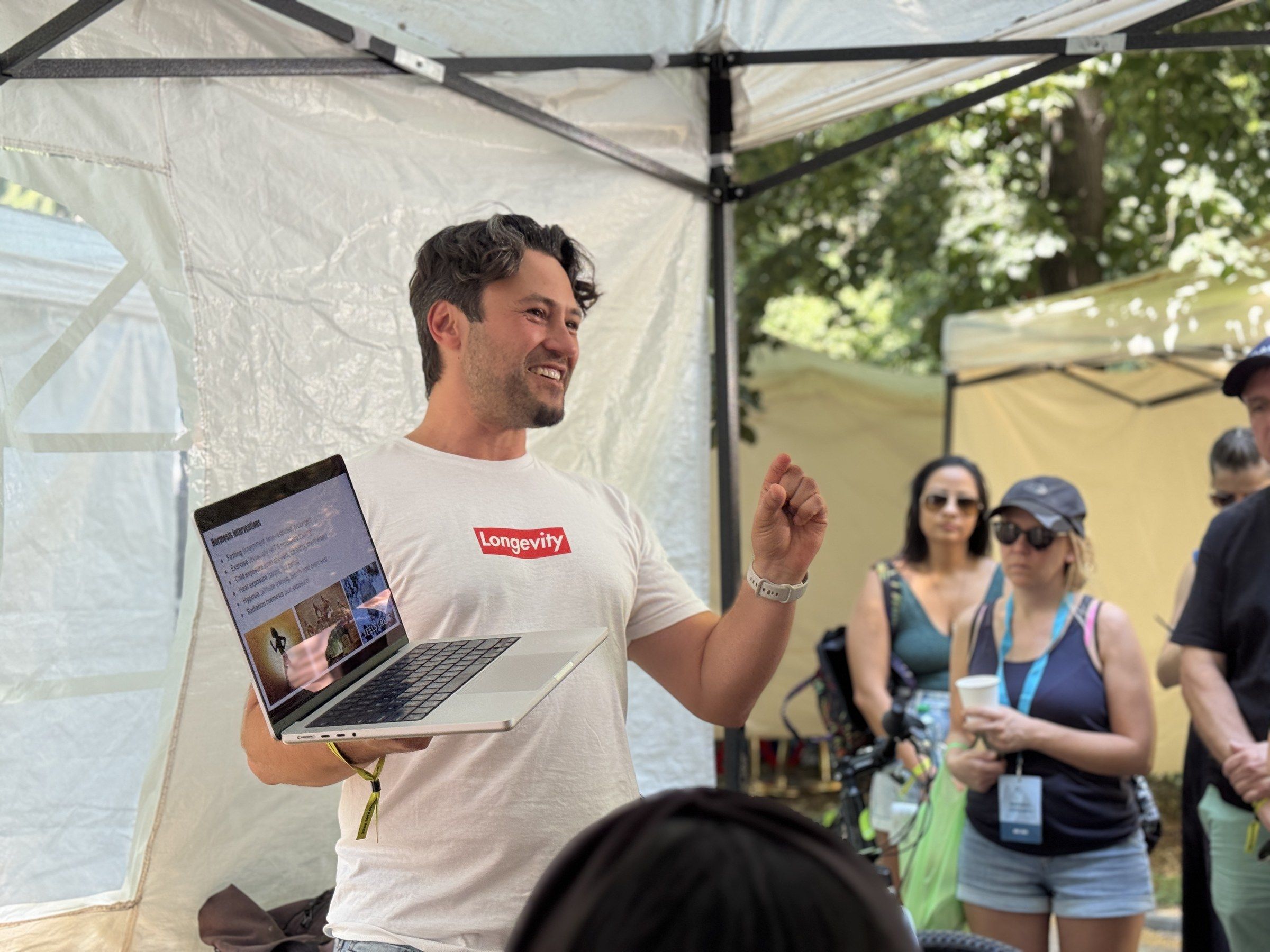 Daniel Duma in a “Longevity” T-shirt presenting from a laptop to a crowd in an outdoor tent